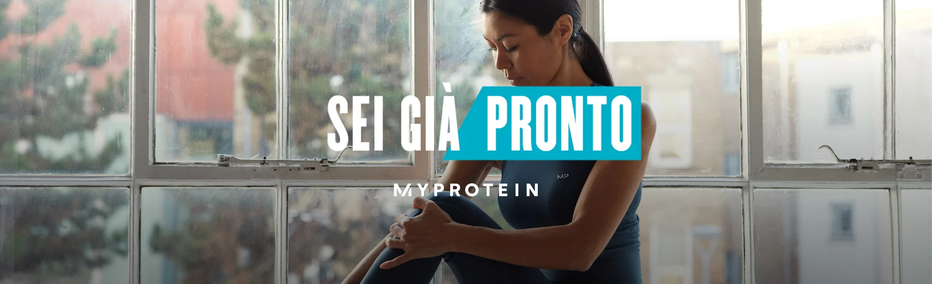 Una donna che indossa abbigliamento da ginnastica MP seduta sul davanzale di una finestra mentre si allunga i muscoli con testo "Sei già pronto - LE MIE PROTEINE" sovrapposto all'immagine.